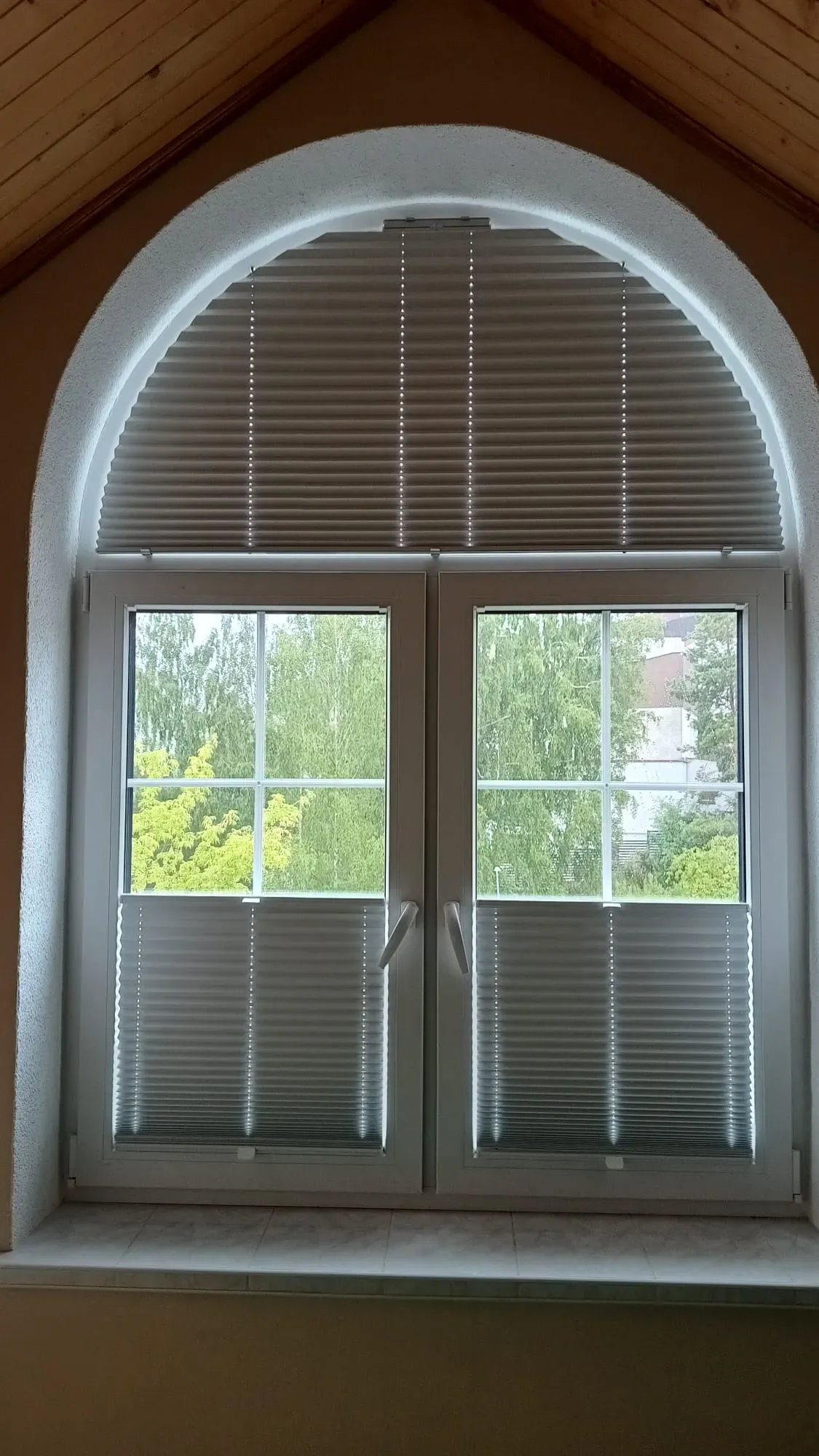 Montaż plis M&G na oknie łukowym w sypialni na poddaszu. Dolne plisy opuszczone do połowy, górna plisa łukowa całkowicie zasłania wnękę okienną.